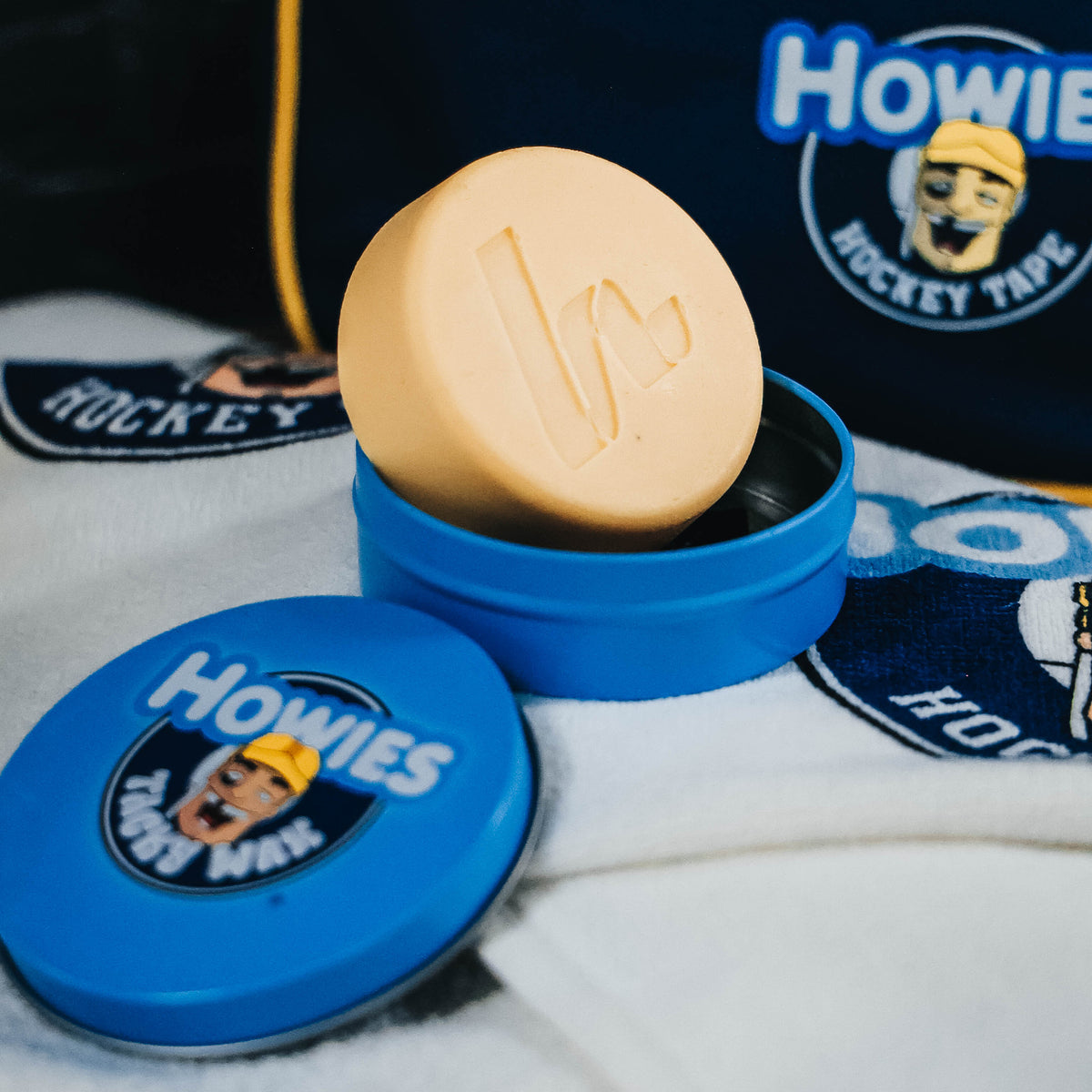 Howies Hockey Tacky Wax tin sitting on a white howies logo bench towel with a blue howies accessory bag in the background 