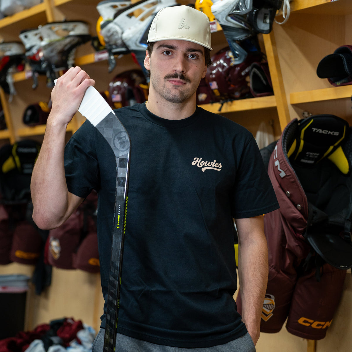 Man holding a hockey stick in a locker room with equipment on shelves.
