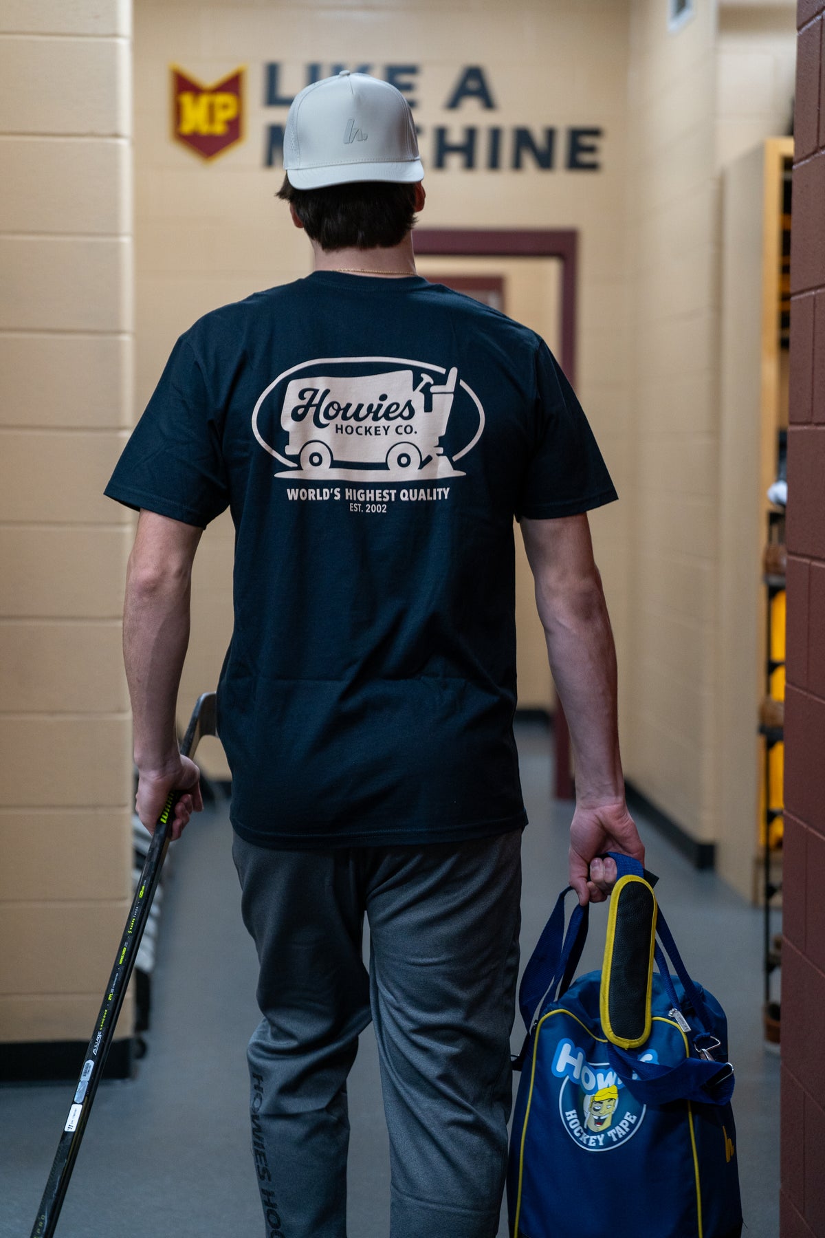 Person wearing a navy blue t-shirt with a logo and text, walking in a hallway.