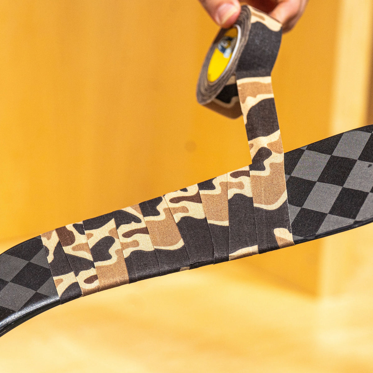 A close-up of a hockey stick being taped with black-and-white checkerboard tape and tan camouflage tape, held by hands in an indoor setting with a wooden wall and floor.