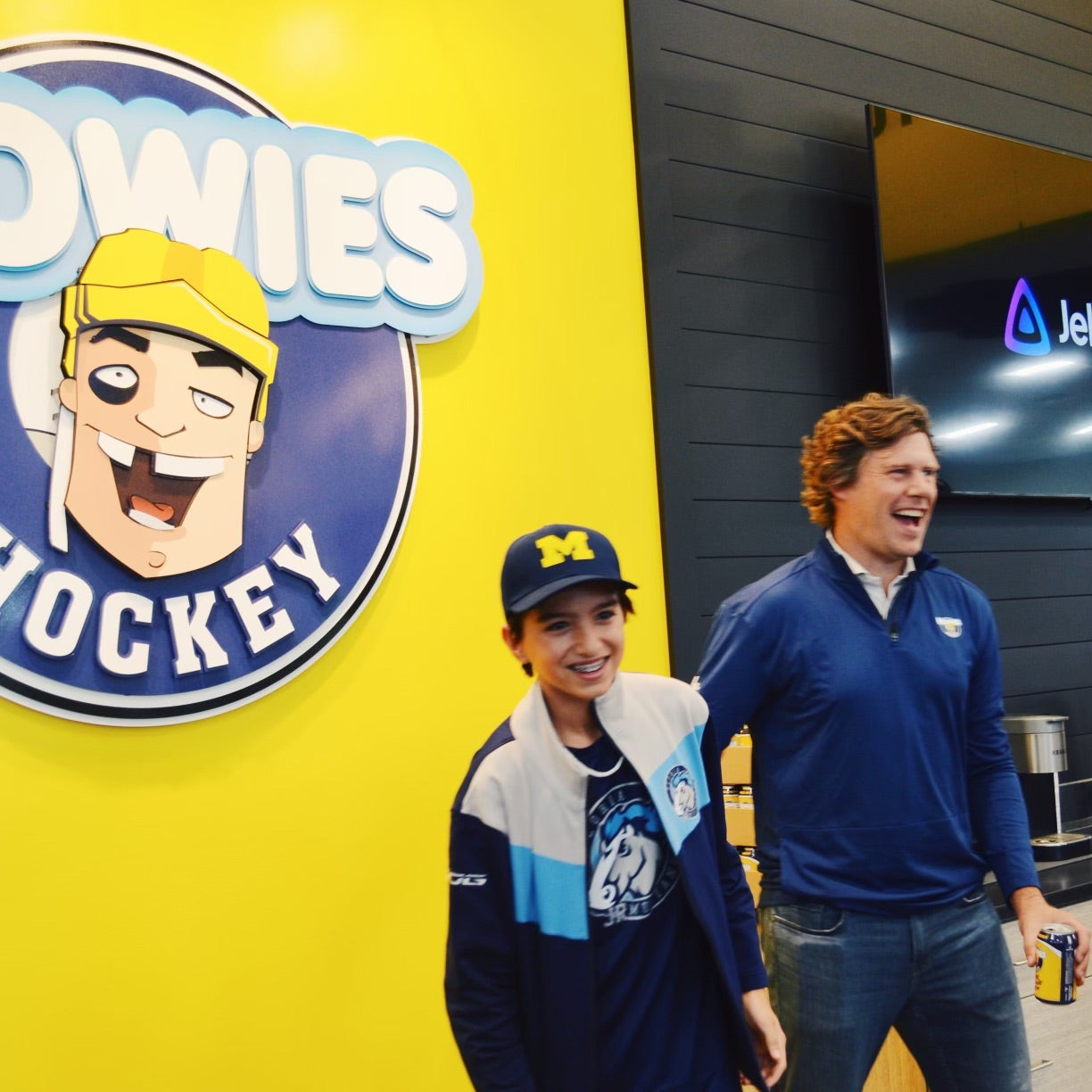 Two people standing in front of a yellow wall with a 'Howies Hockey' logo.