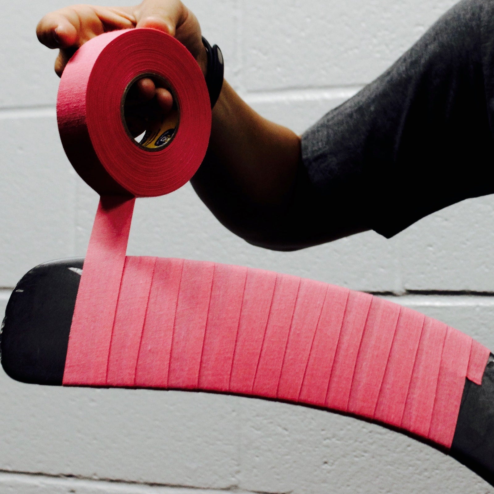 Person applying pink tape to a hockey stick against a white wall