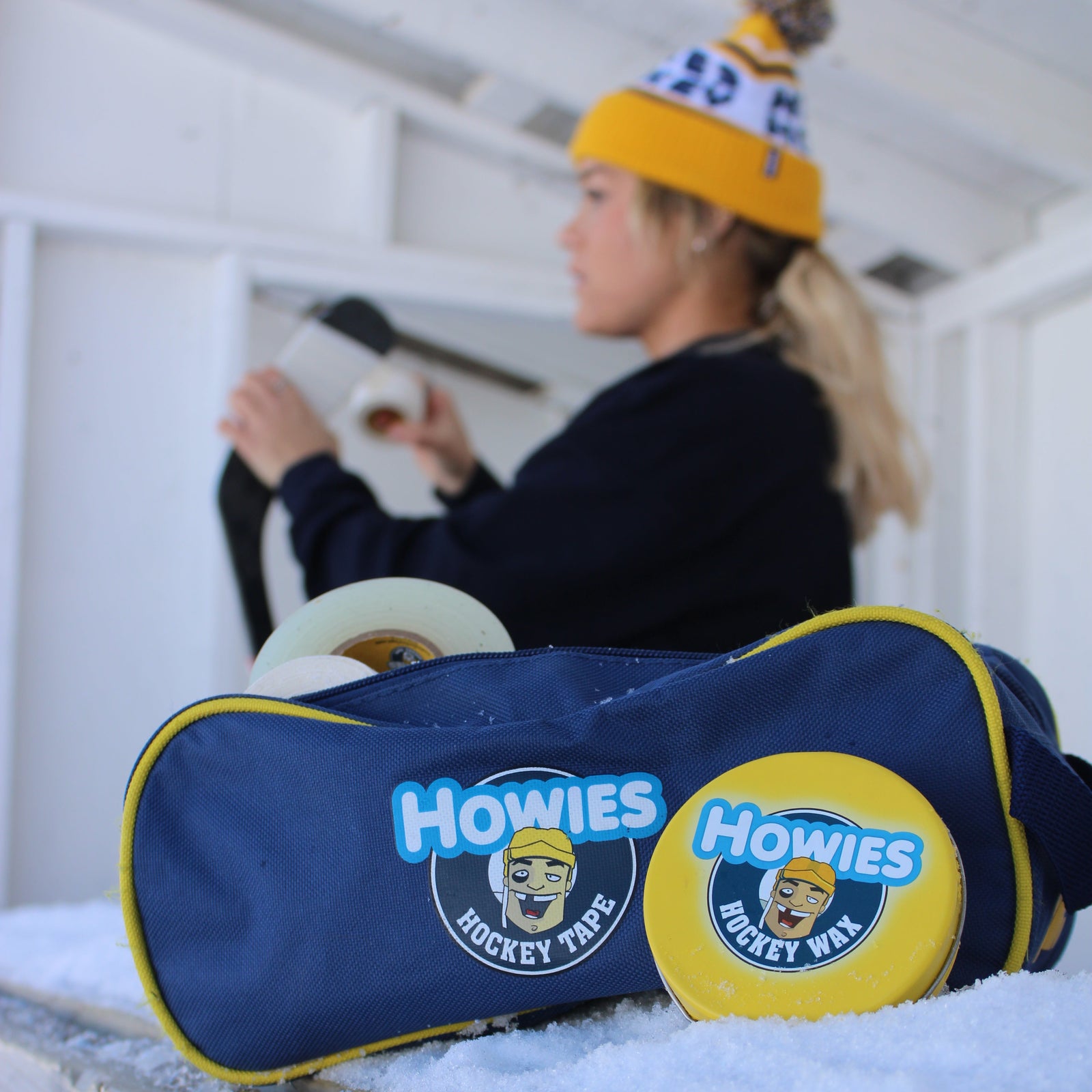 Person taping a hockey stick with white Howies Hockey tape, with a accessory bag filled with tape and wax on a ledge