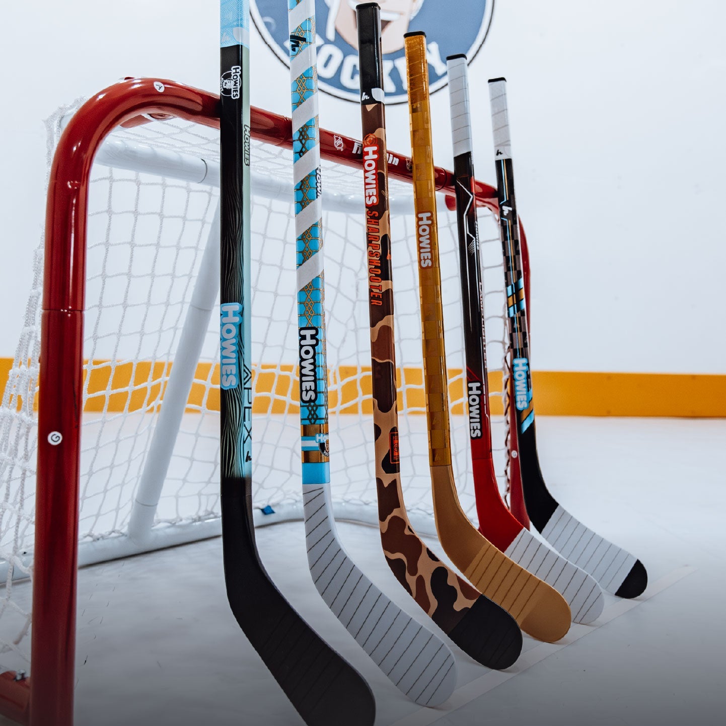 Six hockey sticks with various designs and colors are displayed upright in front of a red hockey goal on an ice rink. A large 'HOMIES' sign is visible in the background.