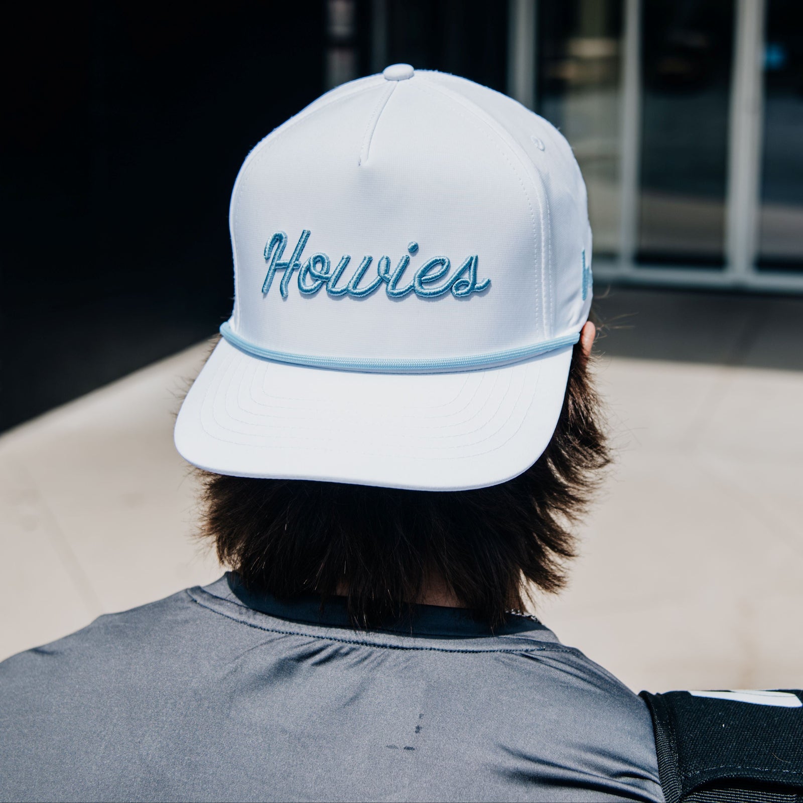 A person viewed from behind wearing a white baseball cap with 'Howies' embroidered in blue script on the front, standing outside near a building entrance.