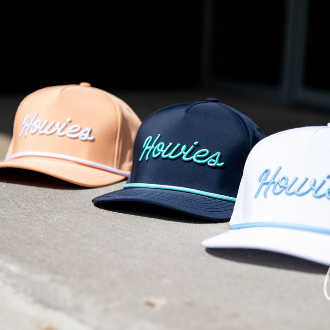 Three snapback caps with 'Howies' embroidered on the front, displayed outdoors on a concrete surface. The caps are peach, navy blue, and white, each with a contrasting light-colored piping detail along the brim and a matching accent stripe on the side.