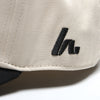 Close-up of the black embroidered Howies "h" logo on the side panel of the cream Clean Sheet Lid